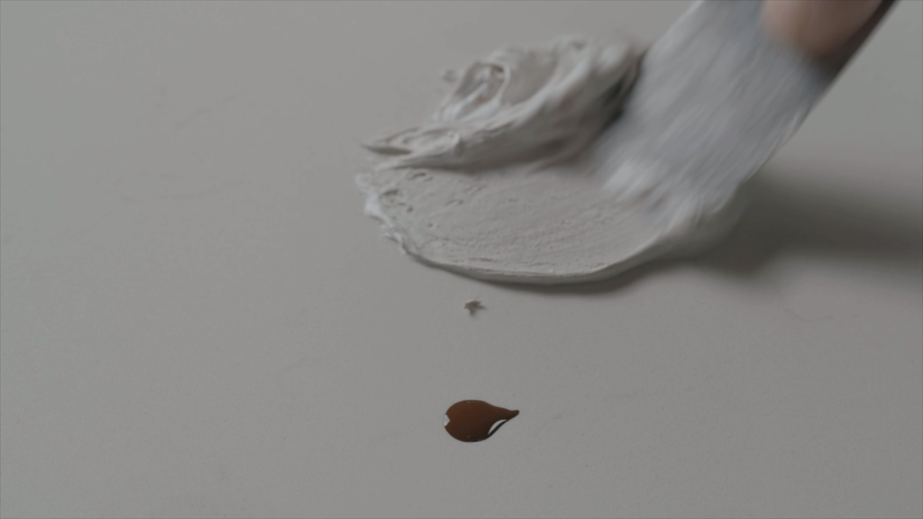 A close-up of a putty knife mixing color pigment into adhesive compound on a porcelain surface for a precise edge repair color-match.