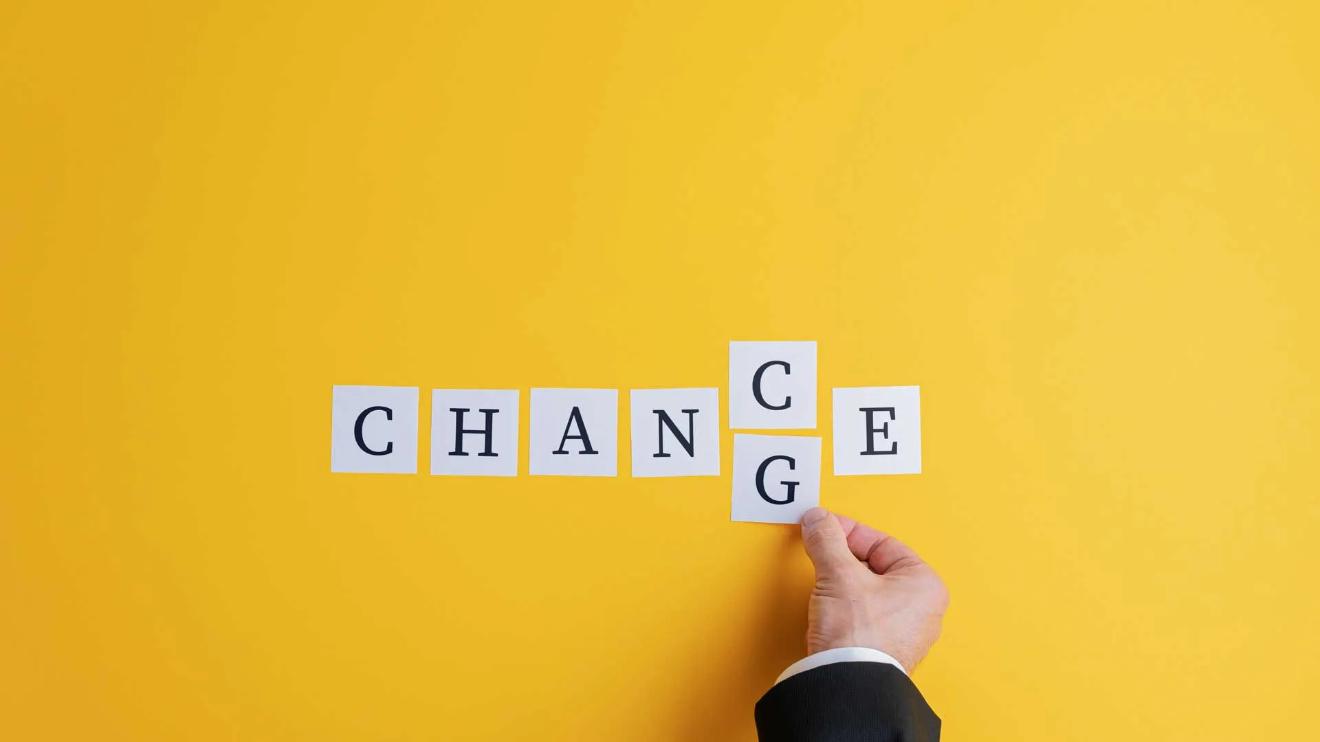 A hand rearranges letter cards spelling “CHANGE” into “CHANCE” against a bright yellow background.