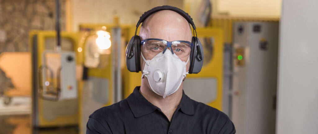 Fabricator wearing protective gear including a respirator mask, safety glasses, and earmuffs, standing confidently in a Caesarstone workshop.