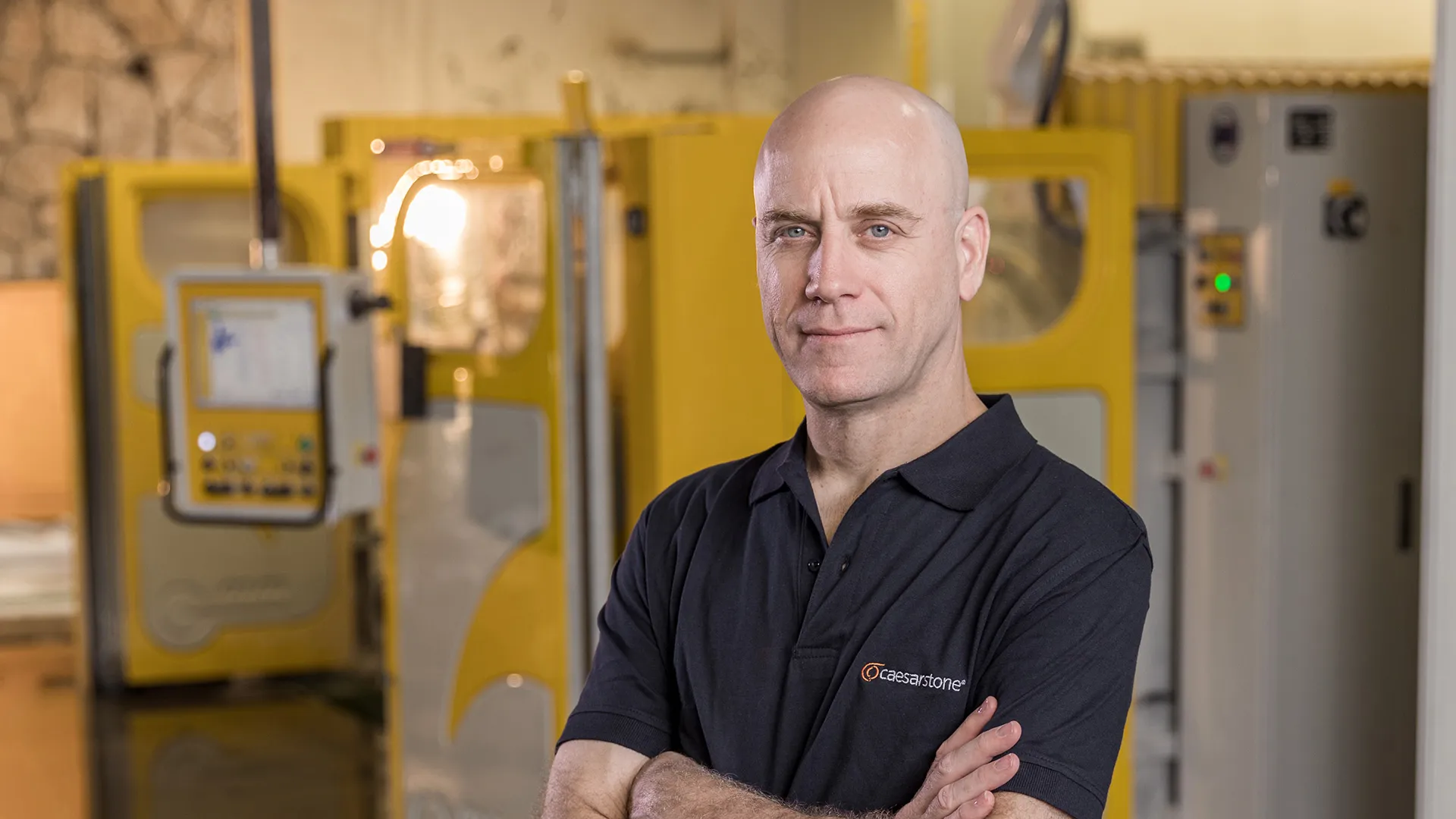 Fabricator at Caesarstone’s Master of Stone Training Center standing confidently in front of machinery, wearing a Caesarstone-branded shirt inside a professional workshop.
