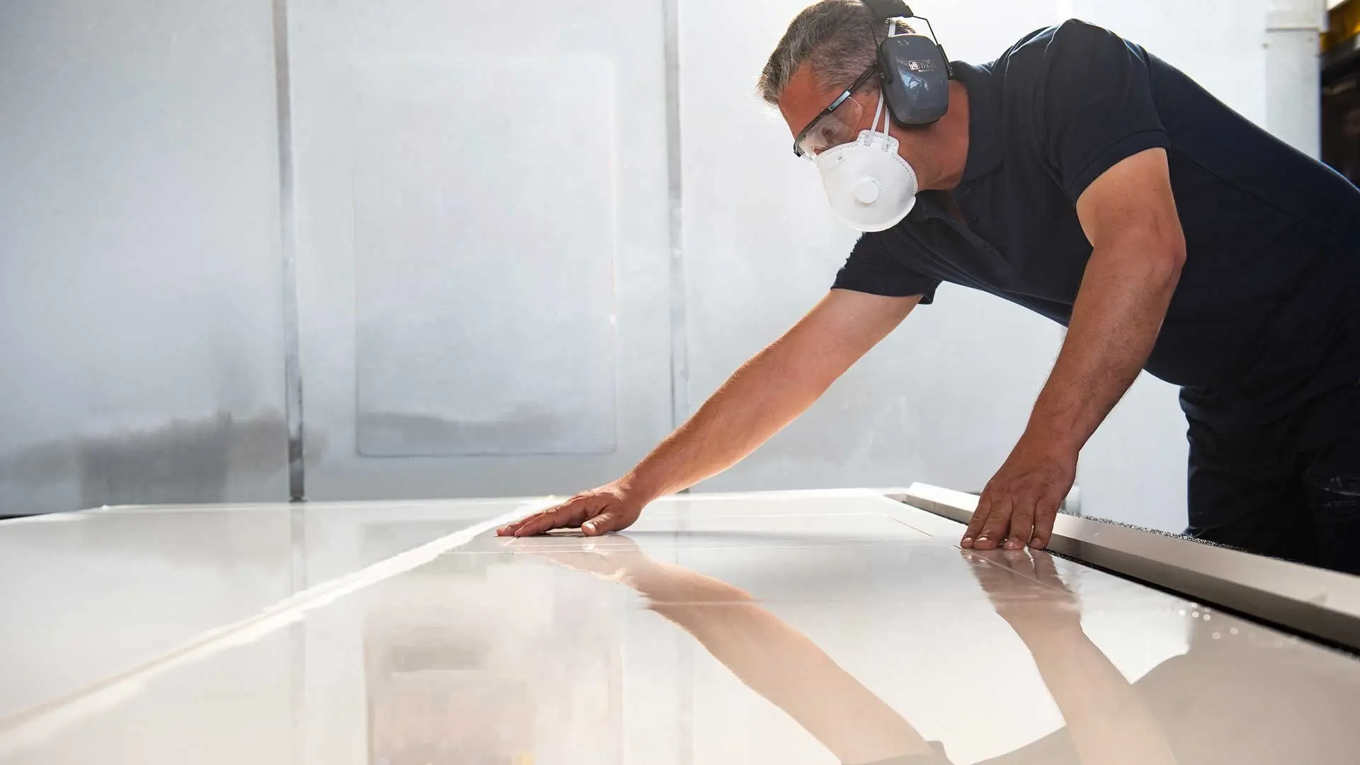 Fabricator wearing protective mask and ear defenders inspecting a polished Caesarstone slab in a workshop, with a “Wear the Right PPE” safety poster in the background.